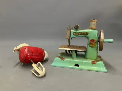 Sale Photo Thumbnail #364: Vintage child's green Sew Master toy sewing machine with floral decals alongside a red toy hand mixer, both showing age-appropriate wear and patina.