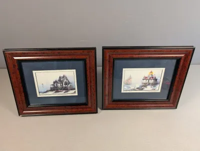 Sale Photo Thumbnail #456: Pair of signed nautical prints featuring coastal structures and lighthouses, displayed in matching burled wood and black composite frames with blue matting.