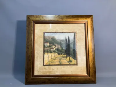 Sale Photo Thumbnail #74: Framed art print depicting a Tuscan landscape with cypress trees and a hillside village, housed in a wide metallic gold-finished wooden frame with double matting.