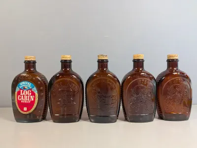 Sale Photo Thumbnail #191: Collection of five vintage 1976 Log Cabin Syrup Bicentennial amber glass flasks with cork stoppers, featuring embossed patriotic designs including the Liberty Bell and Mount Rushmore.