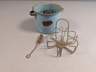 Sale Photo Thumbnail #200: Vintage light blue enamel pot with handles alongside a rusted wire bottle carrier caddy and bottle brush.