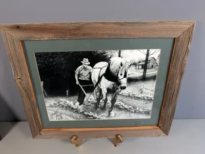 Sale Photo Thumbnail #177: Framed black-and-white print depicting a man plowing a field with a draft horse, housed in a rustic weathered wood frame with a green mat.