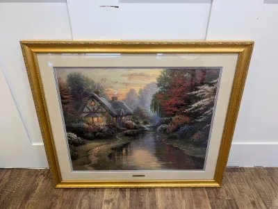 Sale Photo Thumbnail #476: Framed Thomas Kinkade print depicting a cozy cottage by a stream, presented in an ornate gold frame with double matting.
