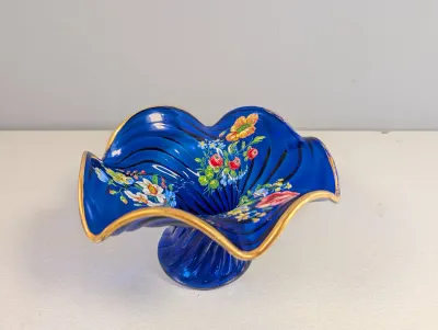Sale Photo Thumbnail #181: Cobalt blue art glass pedestal dish featuring hand-painted floral motifs and a ruffled, gold-rimmed edge.