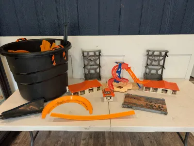 Sale Photo Thumbnail #494: Large assortment of Hot Wheels track pieces, plastic buildings, and Monster Jam playset components, stored in a black bucket and displayed on a table.
