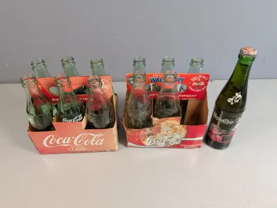 Sale Photo Thumbnail #199: Two cardboard six-pack carriers filled with vintage glass Coca-Cola contour bottles alongside a single green glass Mountain Dew bottle.