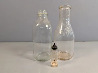 Sale Photo Thumbnail #392: Trio of vintage clear glass bottles, including two milk bottles and one small miniature bottle with a rubber cap, showing light wear.