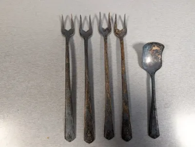Sale Photo Thumbnail #416: Set of four vintage two-prong cocktail forks and one small sugar shell spoon, all showing heavy silver tarnish.