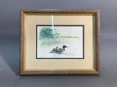 Sale Photo Thumbnail #165: Framed artist proof print of a loon and chick in a lake, signed and dated '87 by Lynda Phillips. Mounted in a textured fabric mat and a wood-tone frame with gold inner trim.