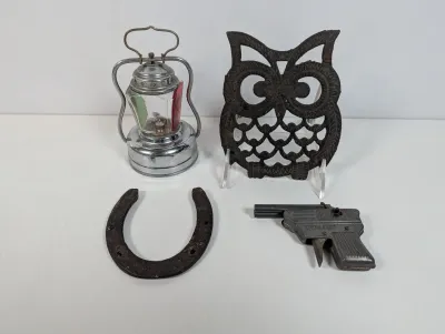 Sale Photo Thumbnail #100: Collection of metal vintage items including a miniature lantern with red and green glass, a cast iron owl trivet, a worn horseshoe, and a metal cap gun.