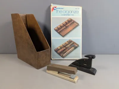 Sale Photo Thumbnail #409: Vintage desk accessories lot including a brown faux-leather magazine holder, two desk staplers, and a boxed Sterling plastic compartment tray.