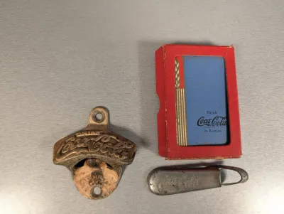 Sale Photo Thumbnail #419: Vintage Coca-Cola collectables including a cast iron wall-mounted bottle opener, a deck of playing cards in its original red window box, and a metallic pocket bottle opener.