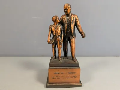 Sale Photo Thumbnail #310: Bronze-tone metal figurine depicting Milton S. Hershey with a young boy, mounted on an inscribed rectangular plinth.