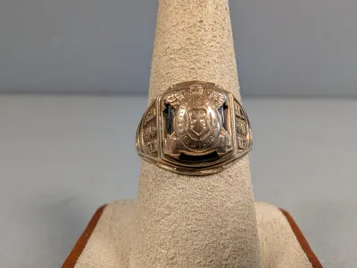 Sale Photo Thumbnail #398: Vintage gold-tone class ring featuring an engraved crest and detailed shoulder accents, displayed on a beige jewelry mandrel.