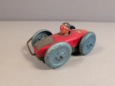 Sale Photo Thumbnail #391: Vintage tin litho rollover race car toy featuring a red body, large metallic grey wheels, and a molded driver with a helmet and goggles.