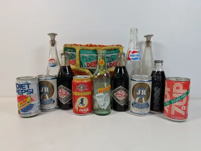 Sale Photo Thumbnail #102: Collection of vintage soda bottles, commemorative cans including J.R. Ewing's Private Stock beer and Super Bowl XXVI Coca-Cola, and a crocheted Mountain Dew bottle holder.