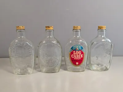 Sale Photo Thumbnail #192: Set of four 1976 Log Cabin Syrup Bicentennial glass commemorative flasks with cork stoppers, featuring embossed historical designs.