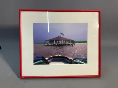 Sale Photo Thumbnail #164: Framed color photograph depicting a floating structure on water from the perspective of a boat, displayed in a bright red frame with off-white matting.