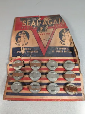 Sale Photo Thumbnail #431: Vintage World War II-era 'Seal-Again' bottle stoppers on the original patriotic display card, featuring a V for Victory logo and eagles. The card shows wear and tearing, and some stoppers exhibit surface rust.