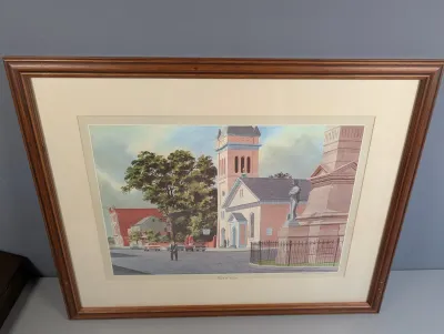 Sale Photo Thumbnail #12: Framed architectural art print depicting a town square with a church, statue, and vintage automobiles, housed under glass in a warm wood frame.