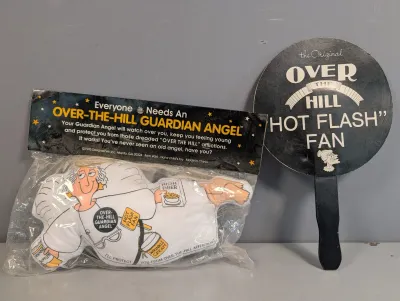 Sale Photo Thumbnail #407: Vintage 1995 Designsense Over-The-Hill Guardian Angel plush doll sealed in original packaging, alongside a matching 'Over The Hill Hot Flash' handheld paddle fan.