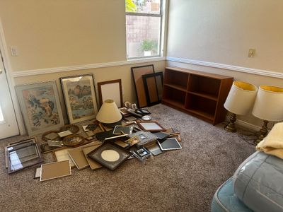 Sale Photo Thumbnail #66: Collection of framed artwork, photo frames, table lamps, and a low wooden bookshelf laid out in a carpeted room.