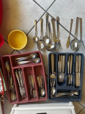 Sale Photo Thumbnail #53: Assorted stainless steel and silver-plated flatware sets housed in pink and blue plastic drawer organizers, accompanied by loose serving utensils and a small yellow ceramic bowl.