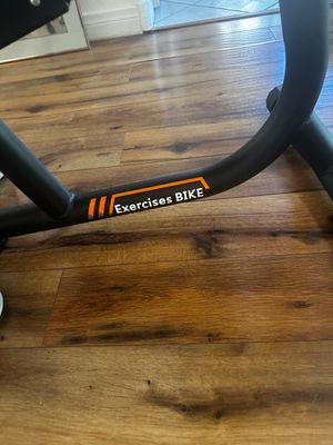 Sale Photo Thumbnail #20: Close-up of a modern black stationary exercise bike frame with orange branding resting on a wood floor.