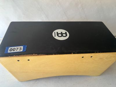 Sale Photo Thumbnail #91: A Meinl brand cajon percussion box instrument featuring a natural wood finish body with a black front playing surface and printed logo. Shows light edge wear from normal use.