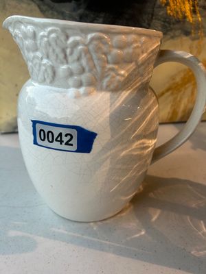 Sale Photo Thumbnail #42: A white ceramic/earthenware pitcher featuring a embossed grape and vine motif around the upper rim and handle. It has an overall crazed/craquelure glazed finish and a sturdy side handle. Appears to be in good pre-owned condition with no visible major chip