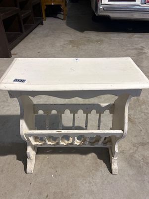 Sale Photo Thumbnail #201: A vintage white-painted wooden side table featuring a built-in lower magazine rack with turned spindle accents and scalloped detail. Shows surface wear, distressing, and paint loss consistent with age and use.