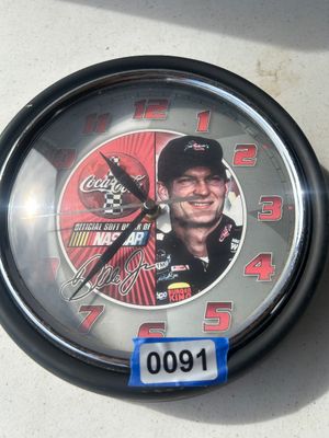 Sale Photo Thumbnail #125: A round wall clock featuring NASCAR driver Dale Earnhardt Jr., Coca-Cola branding, and racing graphics. Frame has a dark casing with chrome trim around the glass dial cover. Measures approximately 10 inches in diameter.