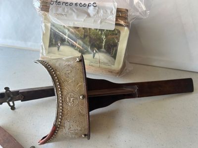 Sale Photo Thumbnail #160: An antique hand-held stereoscope viewer featuring an embossed metal hood with ornate floral and crest details stamped 'MONARCH', a wooden body, folding handle, glass lenses, and slide track. Accompanied by a stack of vintage stereoview/stereograph cards s