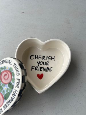 Sale Photo Thumbnail #174: A heart-shaped ceramic trinket box featuring hand-painted floral designs, black and white checkered border details, and inspirational lettering. The inside reads 'CHERISH YOUR FRIENDS', while the lid reads 'THE MOST ESSENTIAL THING FOR HAPPINESS IS THE GI