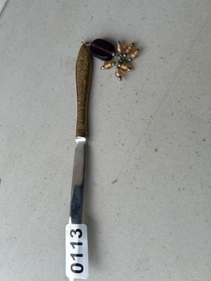 Sale Photo Thumbnail #173: An ornate brass-tone or gold-tone letter opener featuring engraved floral scrollwork on the handle. Attached to the top loop is a decorative dangle charm with purple glass and amber-tone beads. In good vintage condition with minor wear consistent with use