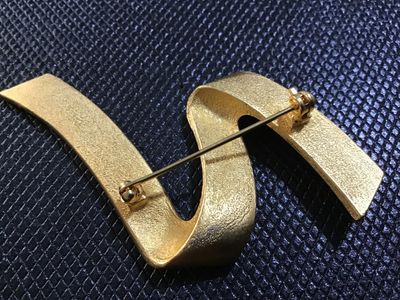 Sale Photo Thumbnail #99: A stylish vintage matte gold-tone brooch featuring a flowing, abstract ribbon or wave design. The reverse side has a textured finish and a standard pin-back closure with a rotary safety clasp. Appears to be in very good vintage condition with light, norma
