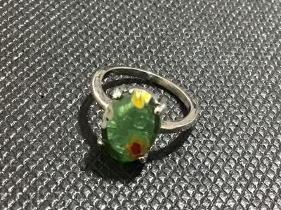 Sale Photo Thumbnail #125: A vintage-style ring featuring an oval green glass cabochon with colorful floral millefiori inclusions. Set in a silver-tone metal band with four double-prongs. Shown on a ring mandrel measuring approximately size 7.5 to 8. The metal displays minor wear a