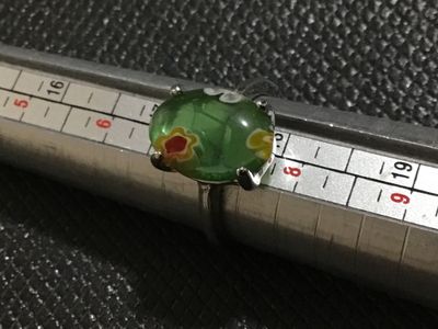 Sale Photo Thumbnail #123: A vintage-style ring featuring an oval green glass cabochon with colorful floral millefiori inclusions. Set in a silver-tone metal band with four double-prongs. Shown on a ring mandrel measuring approximately size 7.5 to 8. The metal displays minor wear a
