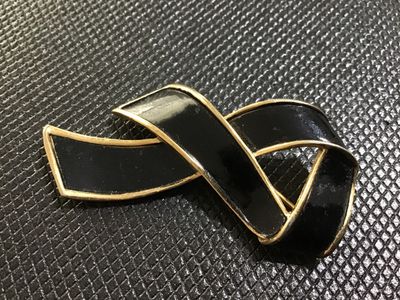 Sale Photo Thumbnail #44: A vintage gold-tone brooch pin featuring a stylish ribbon design with black enamel inlay. Marked 'TRIFARI ©' on the back. Has a textured gold-tone reverse finish with a standard pin stem and roll-over safety clasp. Shows light surface wear consistent wit
