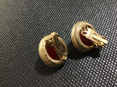 Sale Photo Thumbnail #47: Pair of vintage clip-on earrings featuring round, domed red lucite or resin cabochons set in textured gold-tone metal frames with rope-twist border detailing. Clips show slight vintage wear but are intact and fully functional. Unmarked.