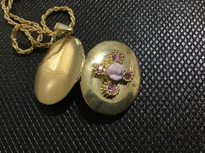 Sale Photo Thumbnail #112: Vintage oval gold-tone locket pendant featuring a central light pink porcelain-style rose detail surrounded by pink rhinestone accents. Suspended on a gold-tone rope chain with a lobster claw clasp. Opens to hold two photos or small mementos. Locket shows