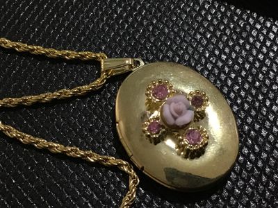 Sale Photo Thumbnail #110: Vintage oval gold-tone locket pendant featuring a central light pink porcelain-style rose detail surrounded by pink rhinestone accents. Suspended on a gold-tone rope chain with a lobster claw clasp. Opens to hold two photos or small mementos. Locket shows