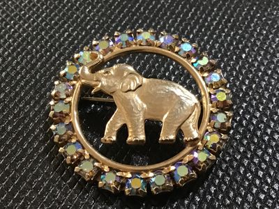 Sale Photo Thumbnail #38: A vintage gold-tone circular brooch by Napier featuring a central textured elephant motif surrounded by a halo of iridescent Aurora Borealis rhinestones. Marked 'J Warner ' on the reverse oval cartouche. Features a roll-over safety clasp. In good vintage 