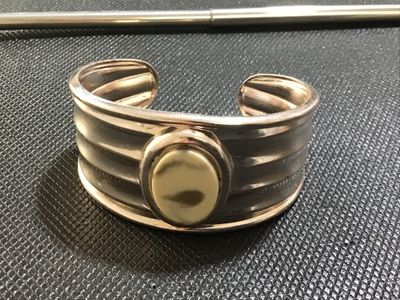 Sale Photo Thumbnail #100: Silver-tone wide ribbed cuff bracelet featuring a central bezel-set oval stone or cabochon accent in a light cream tone. Shows light surface wear and minor tarnishing consistent with pre-owned condition.