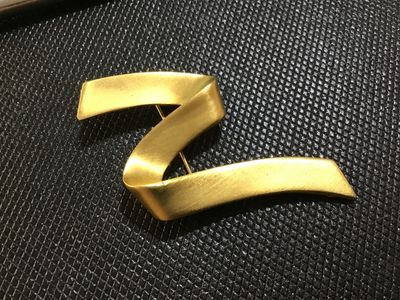 Sale Photo Thumbnail #98: A stylish vintage matte gold-tone brooch featuring a flowing, abstract ribbon or wave design. The reverse side has a textured finish and a standard pin-back closure with a rotary safety clasp. Appears to be in very good vintage condition with light, norma