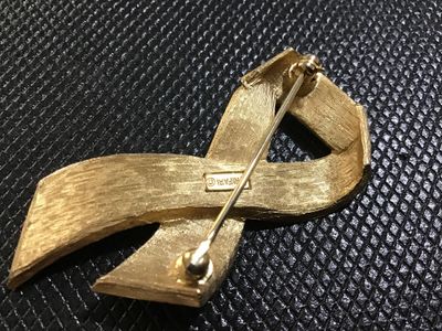 Sale Photo Thumbnail #43: A vintage gold-tone brooch pin featuring a stylish ribbon design with black enamel inlay. Marked 'TRIFARI ©' on the back. Has a textured gold-tone reverse finish with a standard pin stem and roll-over safety clasp. Shows light surface wear consistent wit