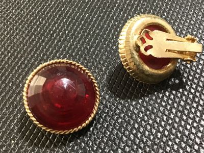 Sale Photo Thumbnail #45: Pair of vintage clip-on earrings featuring round, domed red lucite or resin cabochons set in textured gold-tone metal frames with rope-twist border detailing. Clips show slight vintage wear but are intact and fully functional. Unmarked.