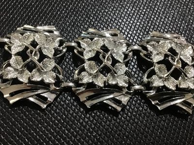 Sale Photo Thumbnail #105: A vintage silver-tone wide link bracelet featuring stylized floral textured leaf motifs within openwork geometric frames. Secured with a fold-over clasp. Appears to be mid-20th century costume jewelry in good condition with minor surface wear visible on t