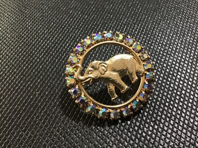 Sale Photo Thumbnail #36: A vintage gold-tone circular brooch by Napier featuring a central textured elephant motif surrounded by a halo of iridescent Aurora Borealis rhinestones. Marked 'J Warner ' on the reverse oval cartouche. Features a roll-over safety clasp. In good vintage 