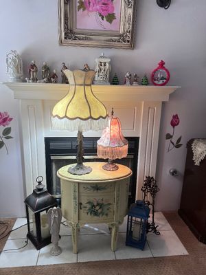 Sale Photo Thumbnail #13: Hand-painted vintage nightstand with floral motifs, displaying two lit table lamps featuring fringed bell shades in front of a white fireplace mantle.